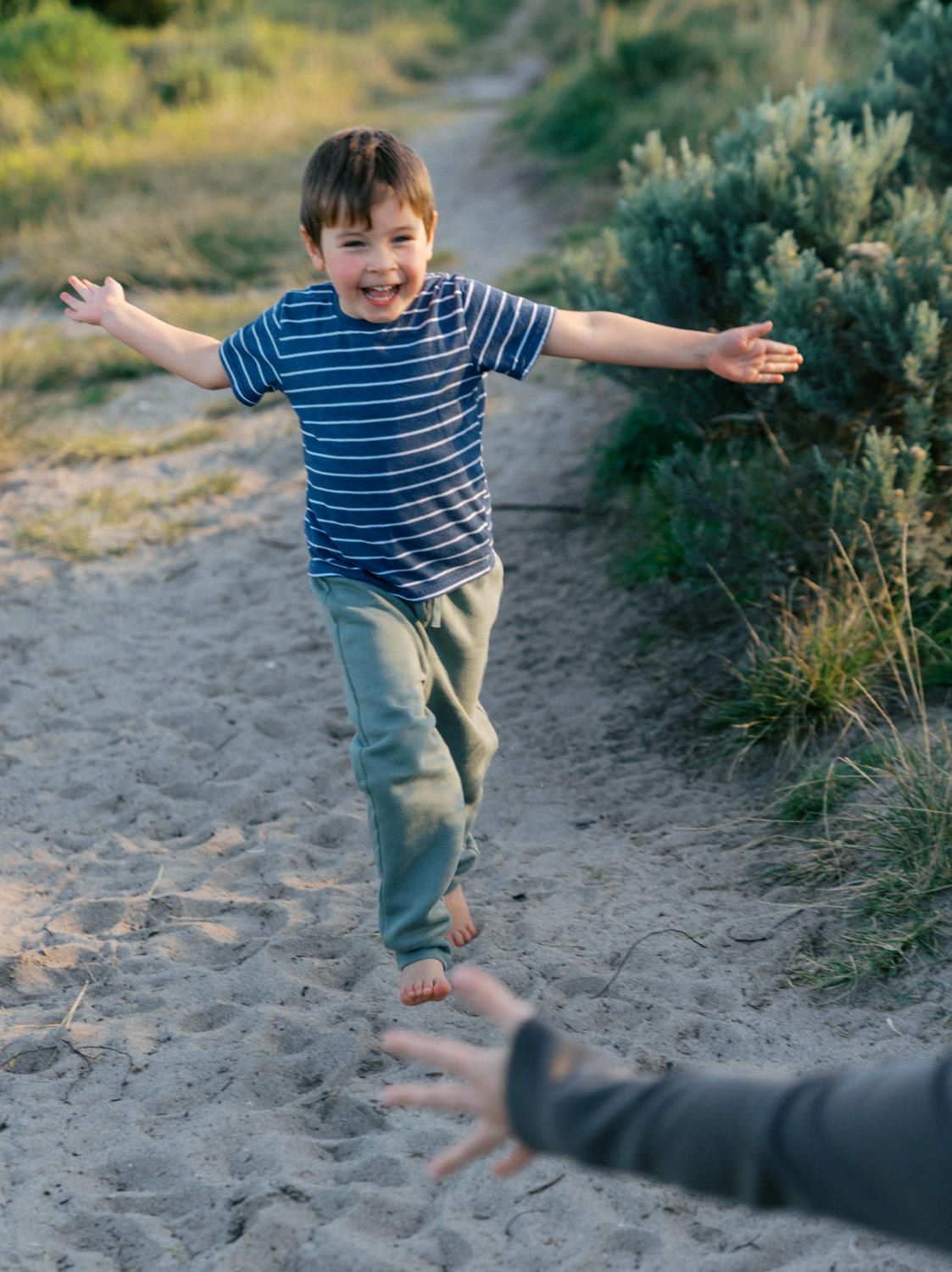 A joyful young boy in a striped shirt runs with arms outstretched on a sandy beach path.