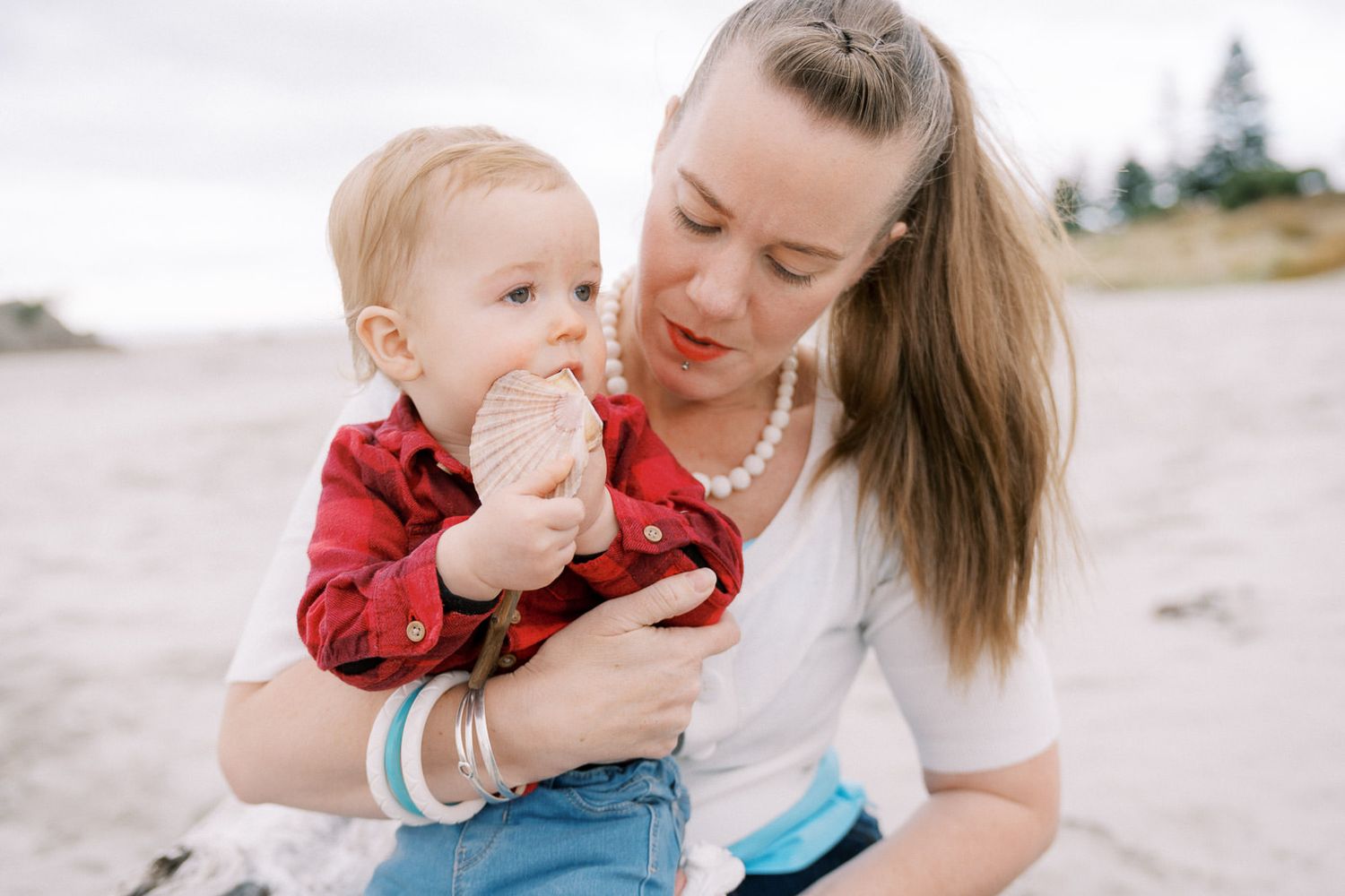 A mother holds her baby on a beach, smiling as the infant chews on a snack, both dressed casually in a serene outdoor setting.