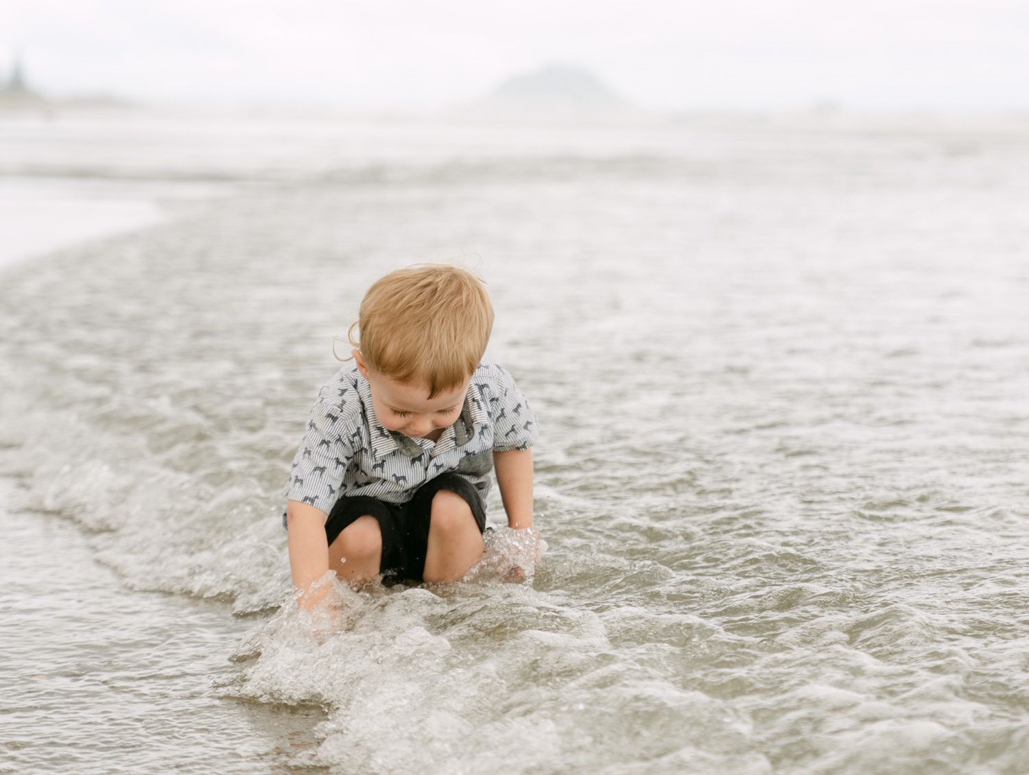 Young boy playing in ocean waves on a sandy beach, wearing a patterned shirt and shorts on a cloudy day.