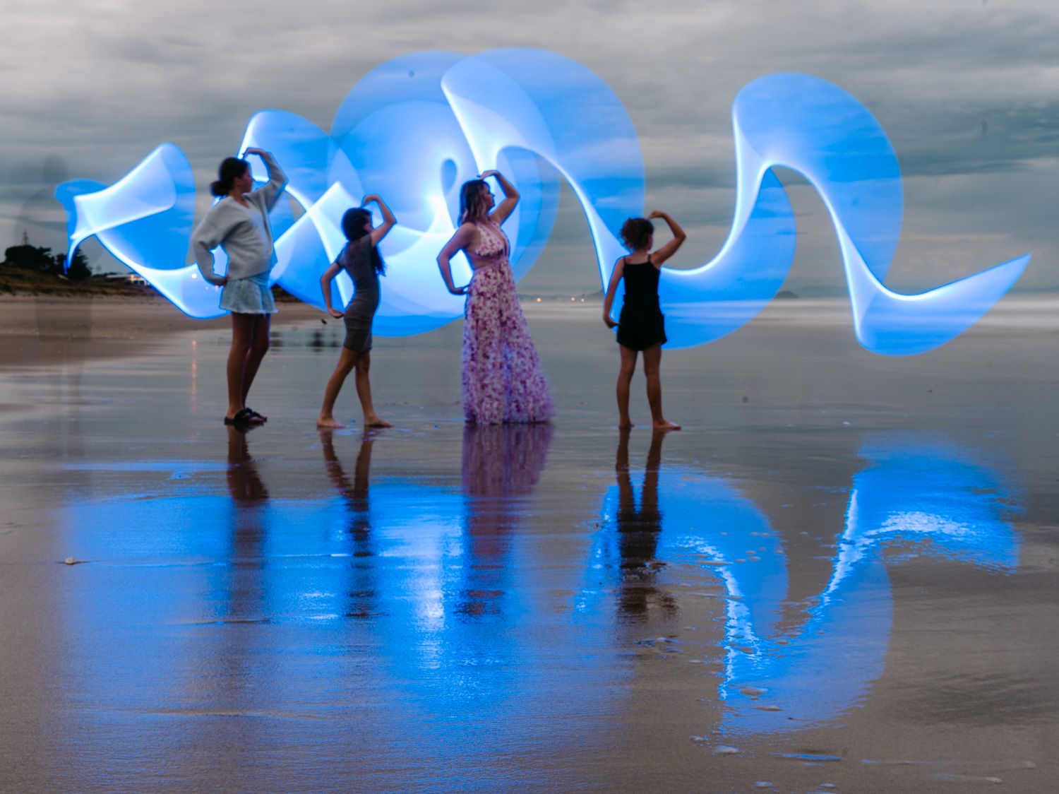 People performing light painting art on a reflective beach at dusk with glowing blue light trails.