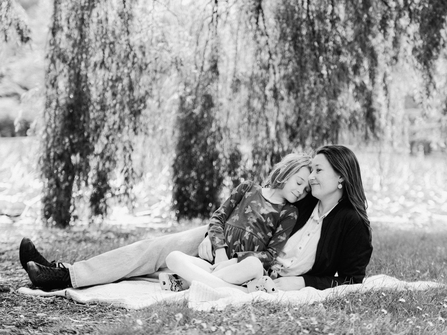 Black and white photo of two women relaxing on a blanket under a weeping willow tree outdoors.