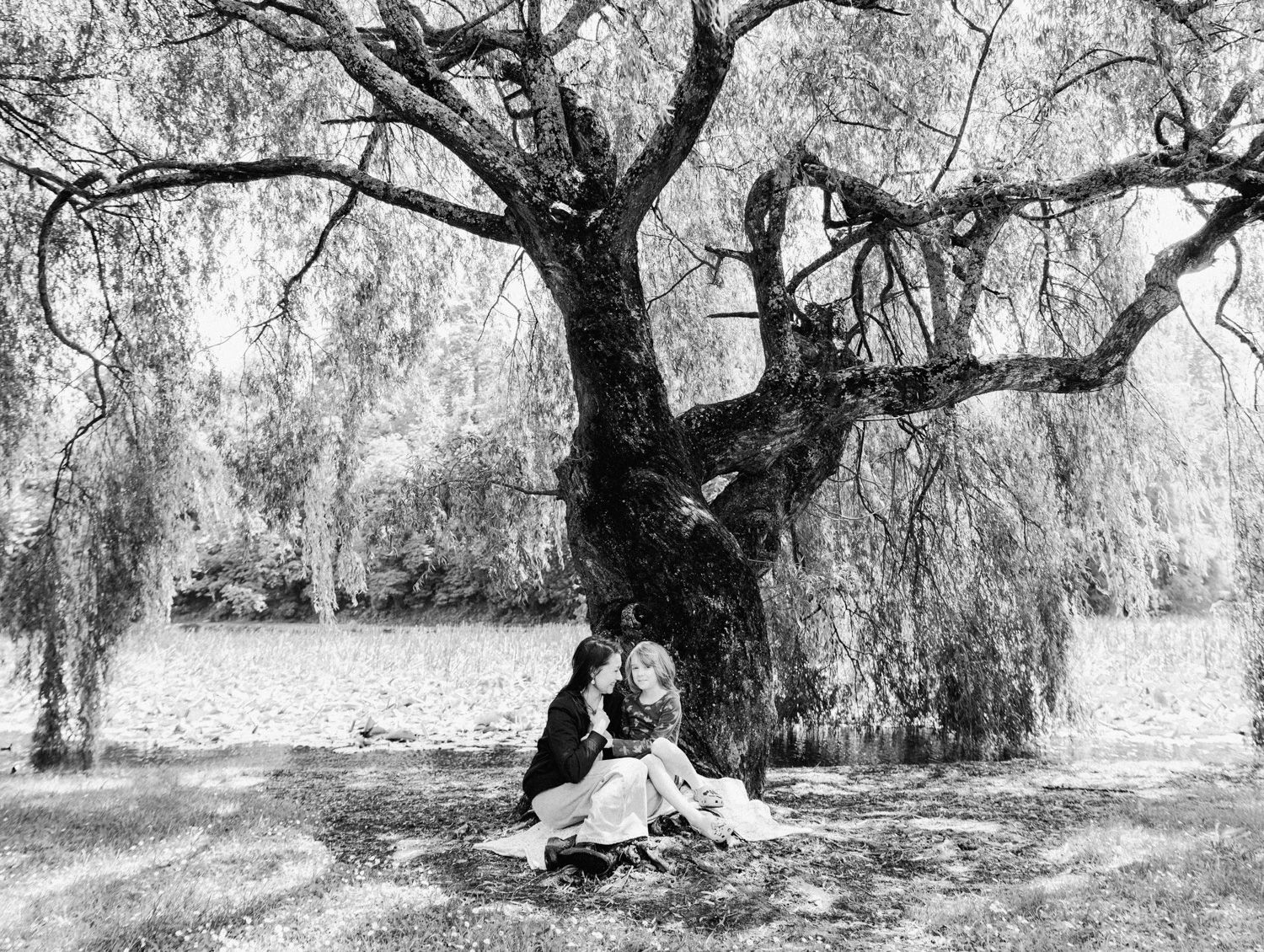 Black and white photo of a child sitting under a large weeping willow tree near a lake.