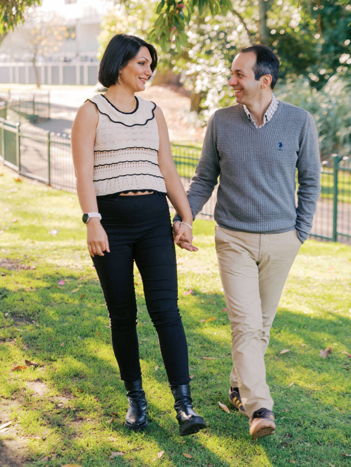 Couple holding hands and smiling at each other while walking through a green park on a sunny day.