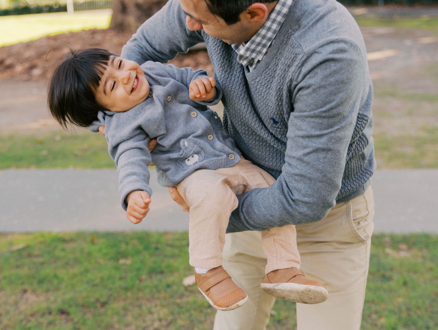 A father joyfully lifts his laughing toddler outdoors in a park, both wearing cozy sweaters and casual pants.