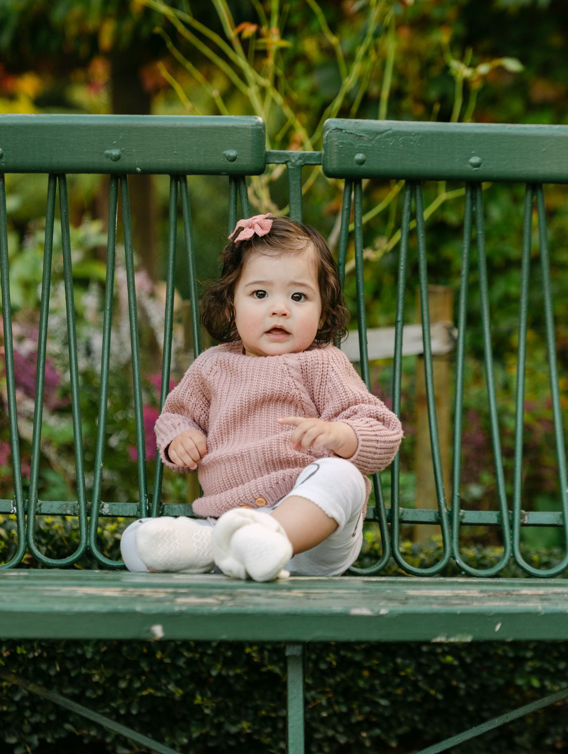 A baby girl in a pink knit sweater sits on a green metal bench surrounded by lush garden greenery.
