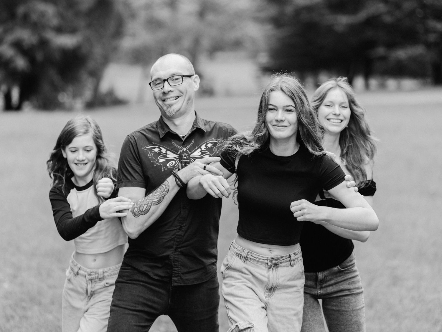 Black and white family photo of a bald man and three teenage girls laughing and posing together outdoors.