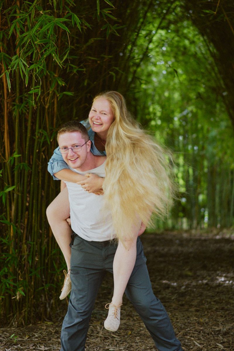 A laughing couple enjoys a piggyback ride in a lush bamboo forest, her long blonde hair flowing freely.