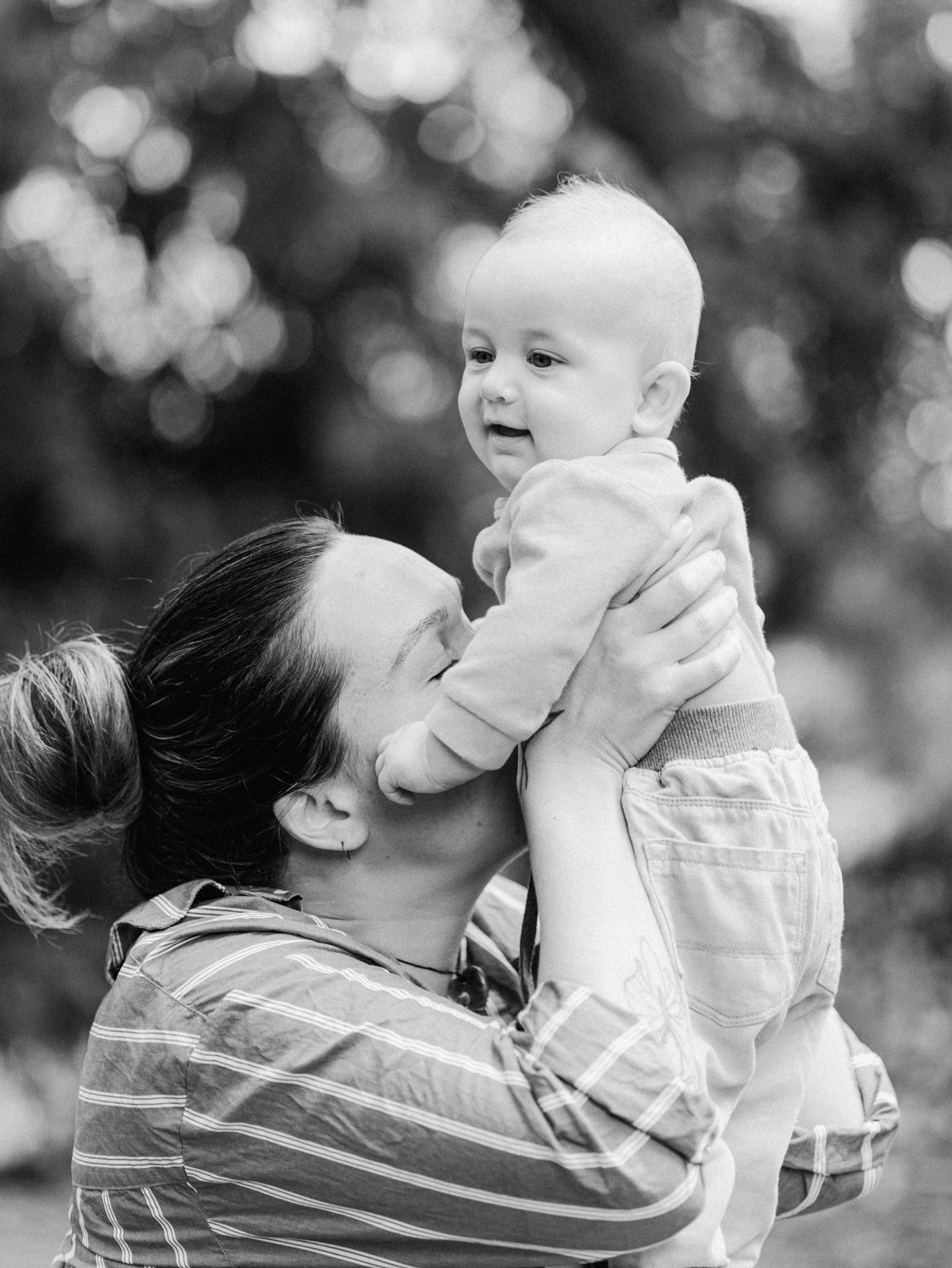 Black and white photo of a parent lovingly lifting a smiling baby outdoors with bokeh background.