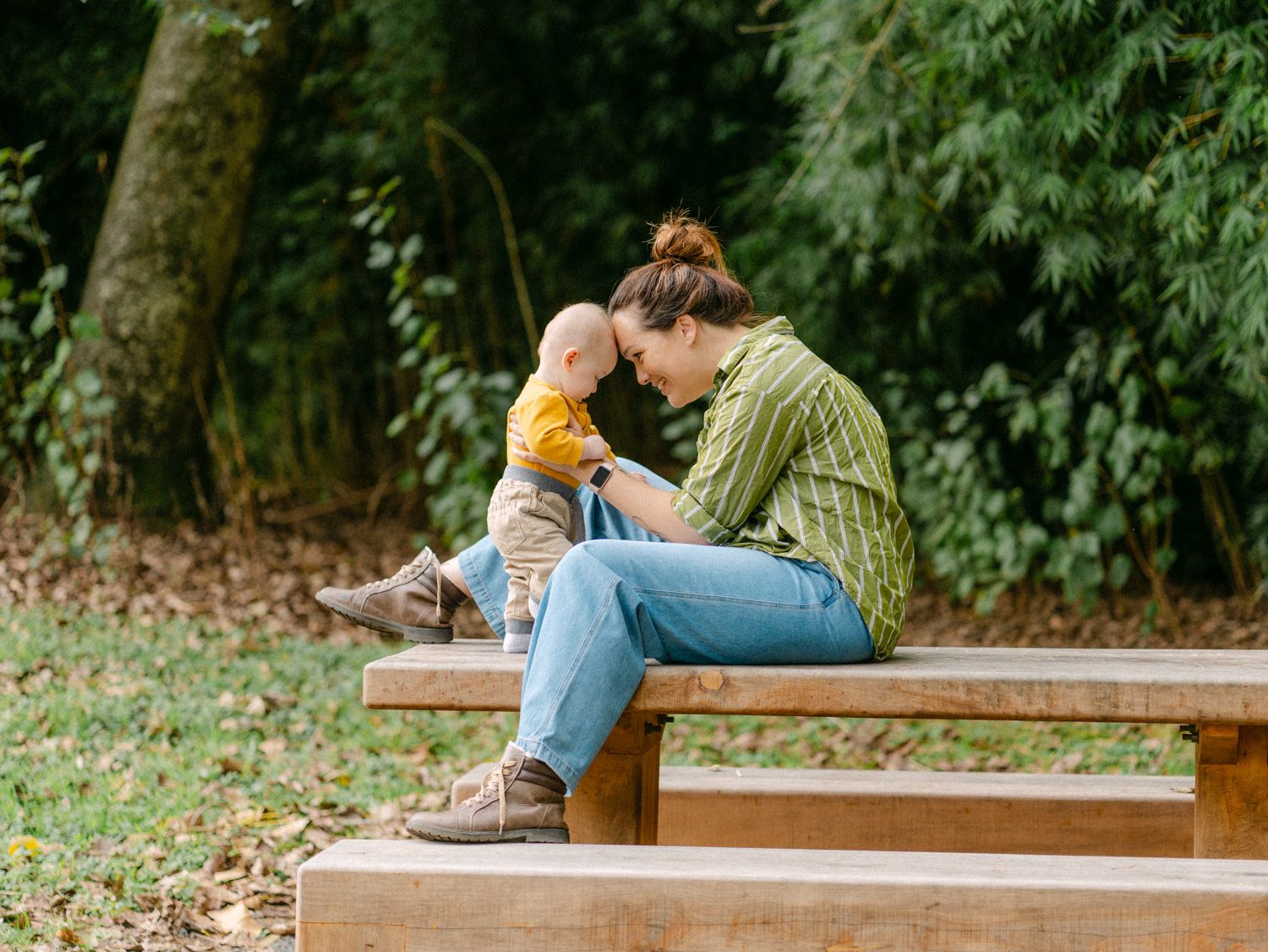 A mother sits on a wooden bench in a park, tenderly touching foreheads with her baby dressed in yellow.