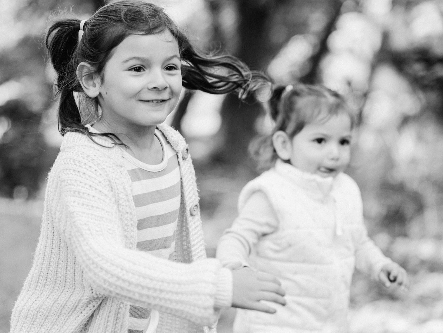 Black and white photo of two young girls playing outdoors, older girl smiling in cardigan, toddler in white vest behind her.