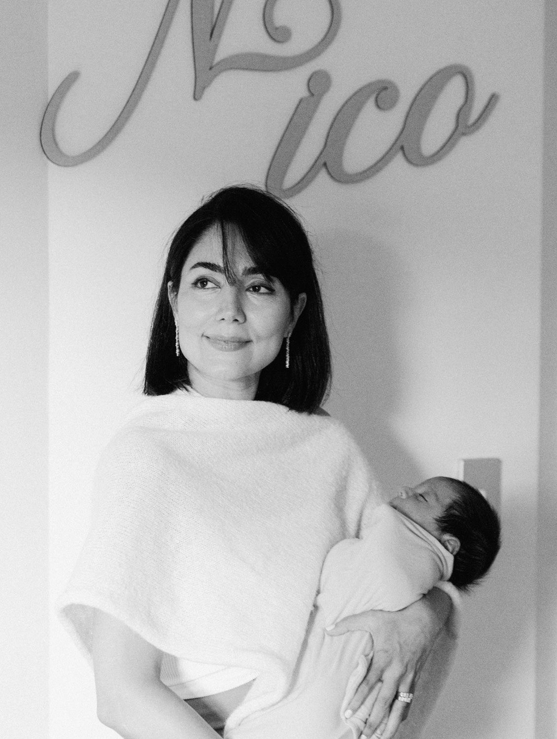 Black and white photo of a mother holding her swaddled newborn beneath a wooden name sign reading Nico.