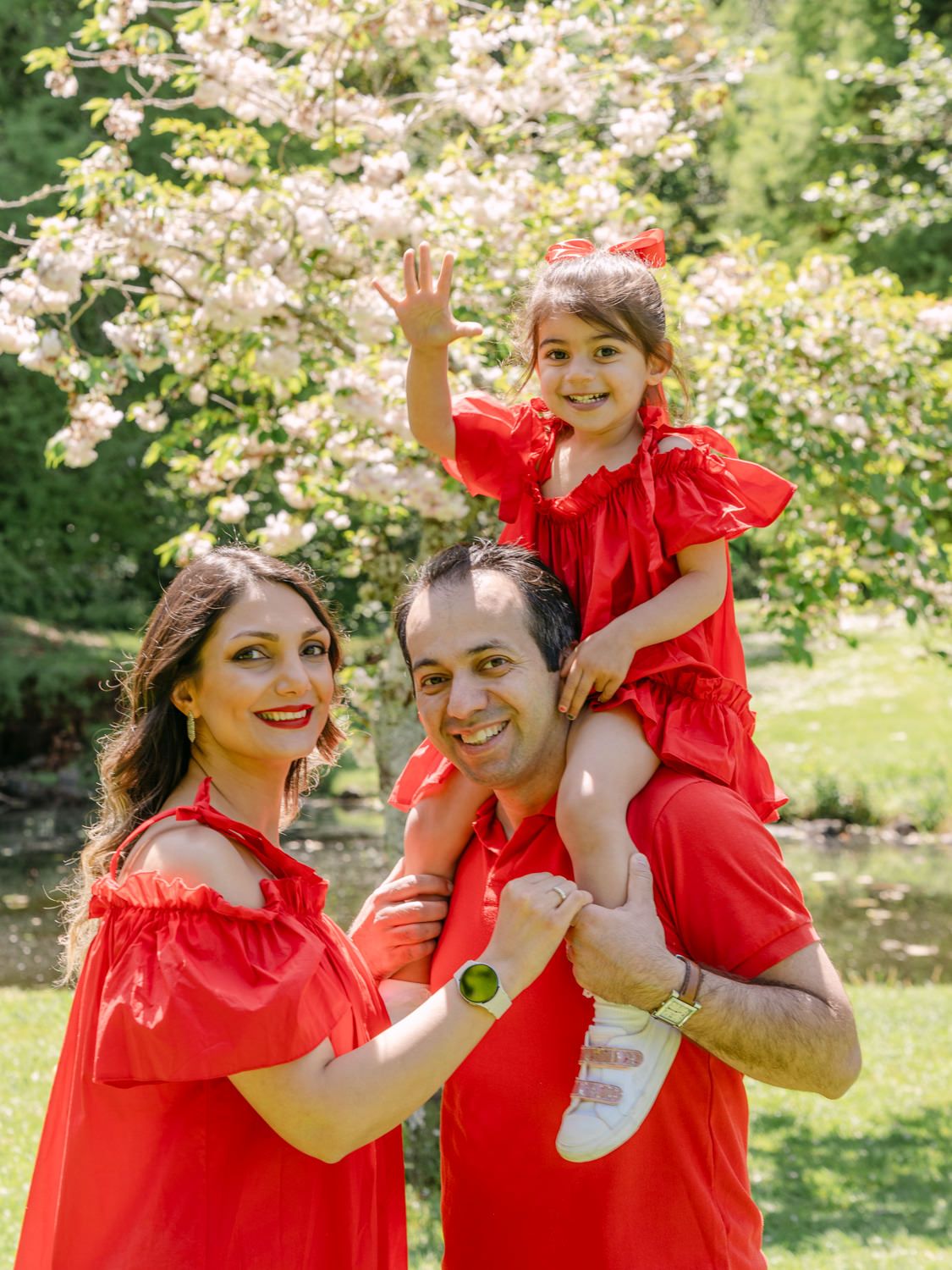 A joyful family in matching red outfits poses outdoors, with a little girl sitting on her father's shoulders waving.