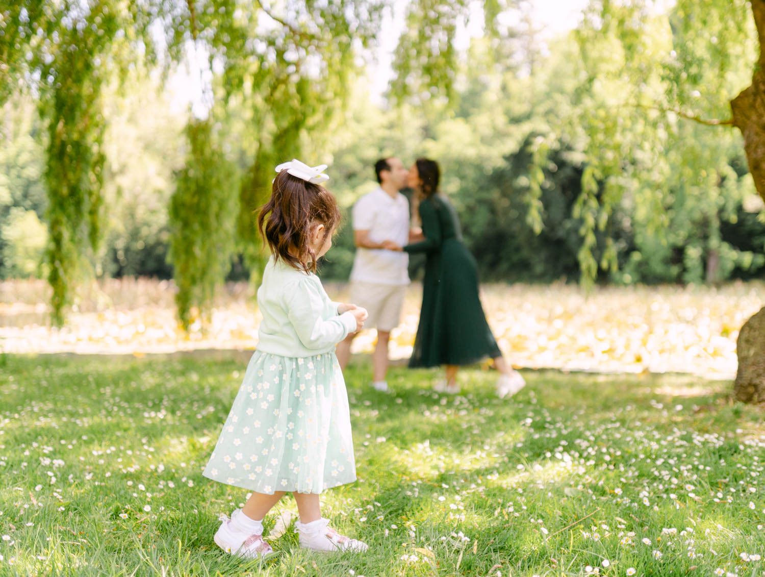 Young girl in mint dress watches parents embrace under weeping willow tree in sunny green park.
