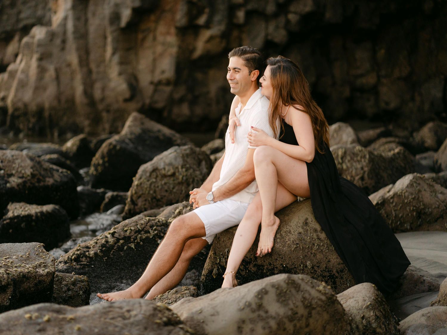 Romantic couple sitting together on coastal rocks by the ocean, woman in black dress embracing man in white shorts.
