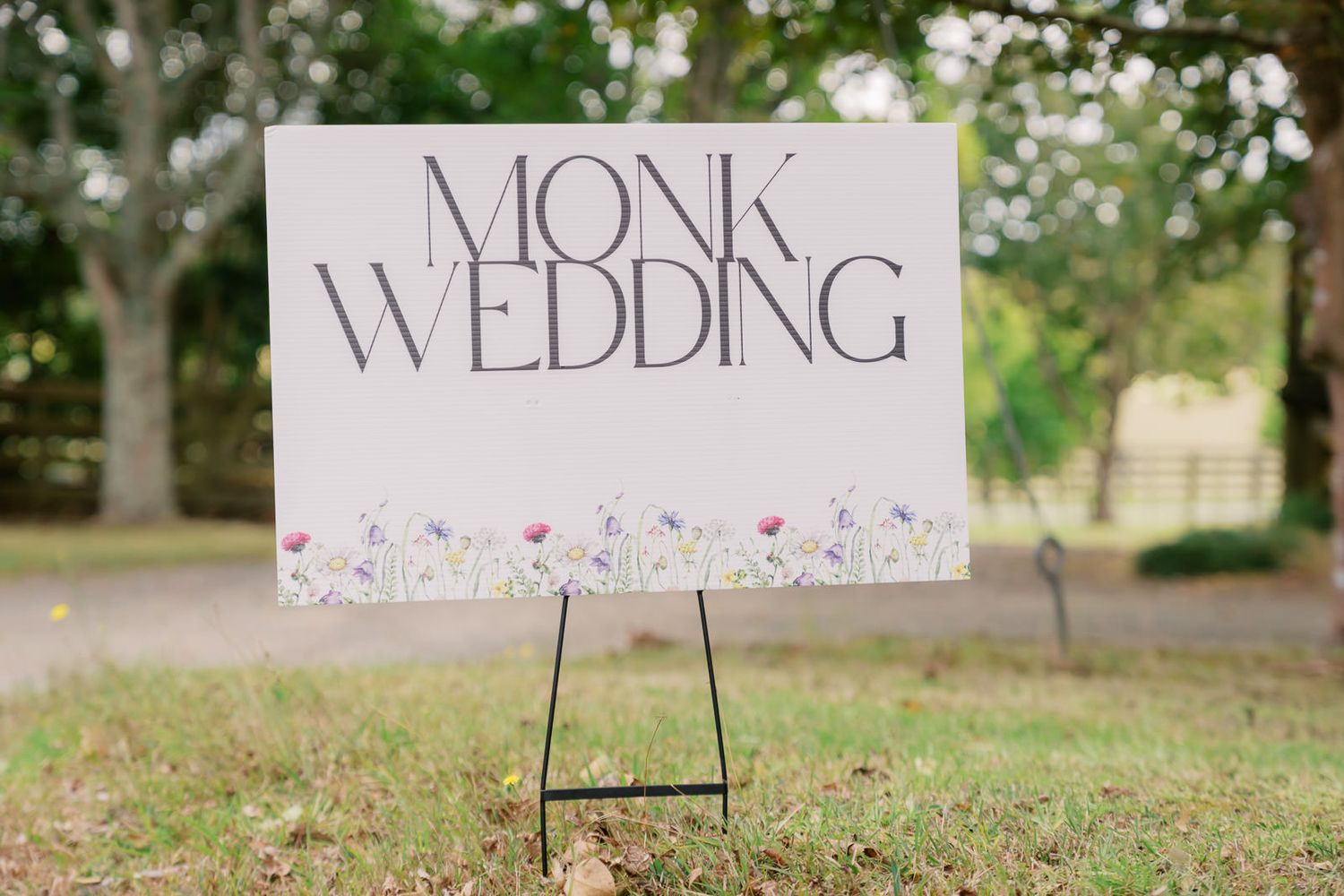 Elegant outdoor wedding welcome sign reading Monk Wedding with wildflower border displayed on black easel stand.