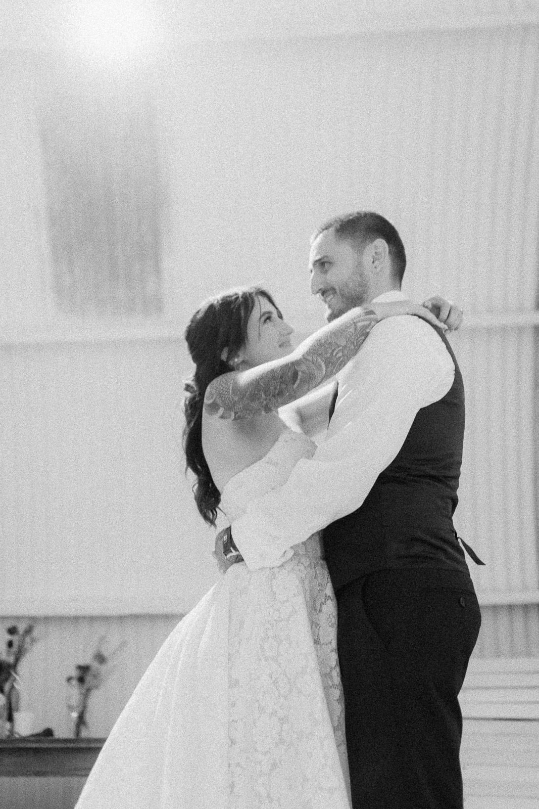 Black and white photo of a bride and groom sharing their first dance, gazing at each other lovingly.