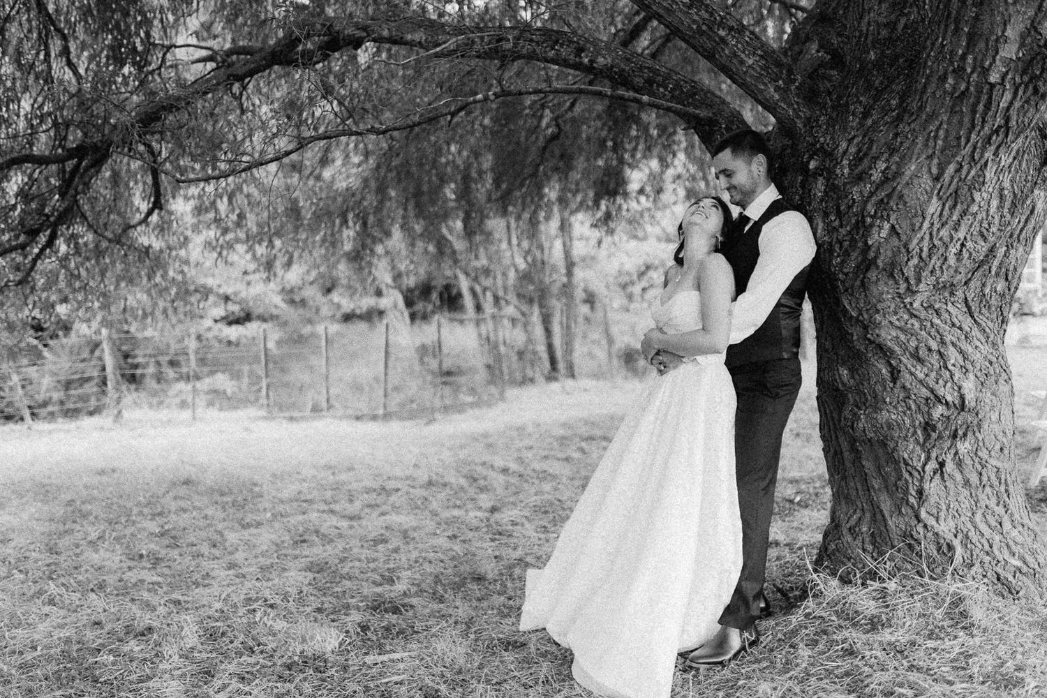 Black and white photo of a bride and groom embracing under a large tree in a lush outdoor setting.