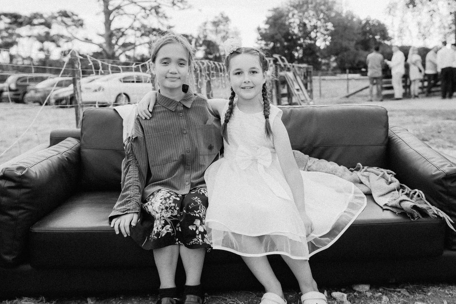 Two young girls in formal attire smile together on a black sofa at an outdoor wedding celebration.