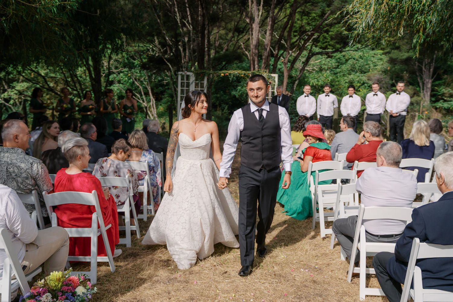 Bride and groom walk hand in hand down outdoor wedding aisle surrounded by seated guests and lush green trees.