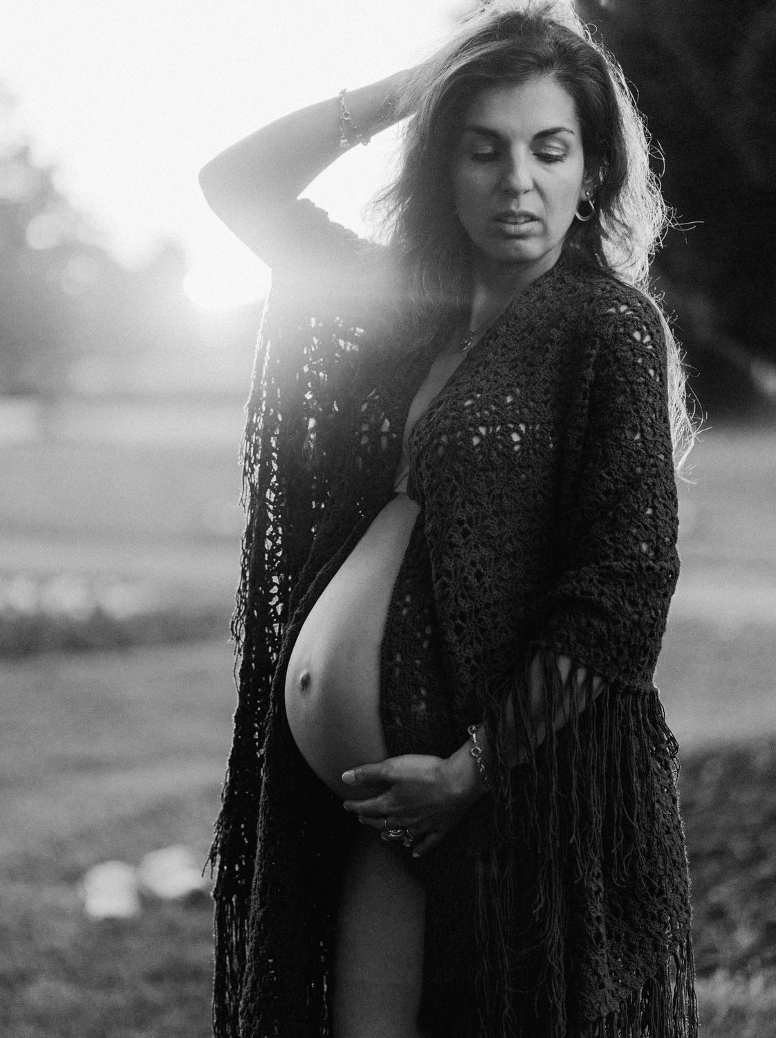 Black and white maternity photo of a pregnant woman in a lace cardigan, cradling her bare belly at sunset outdoors.