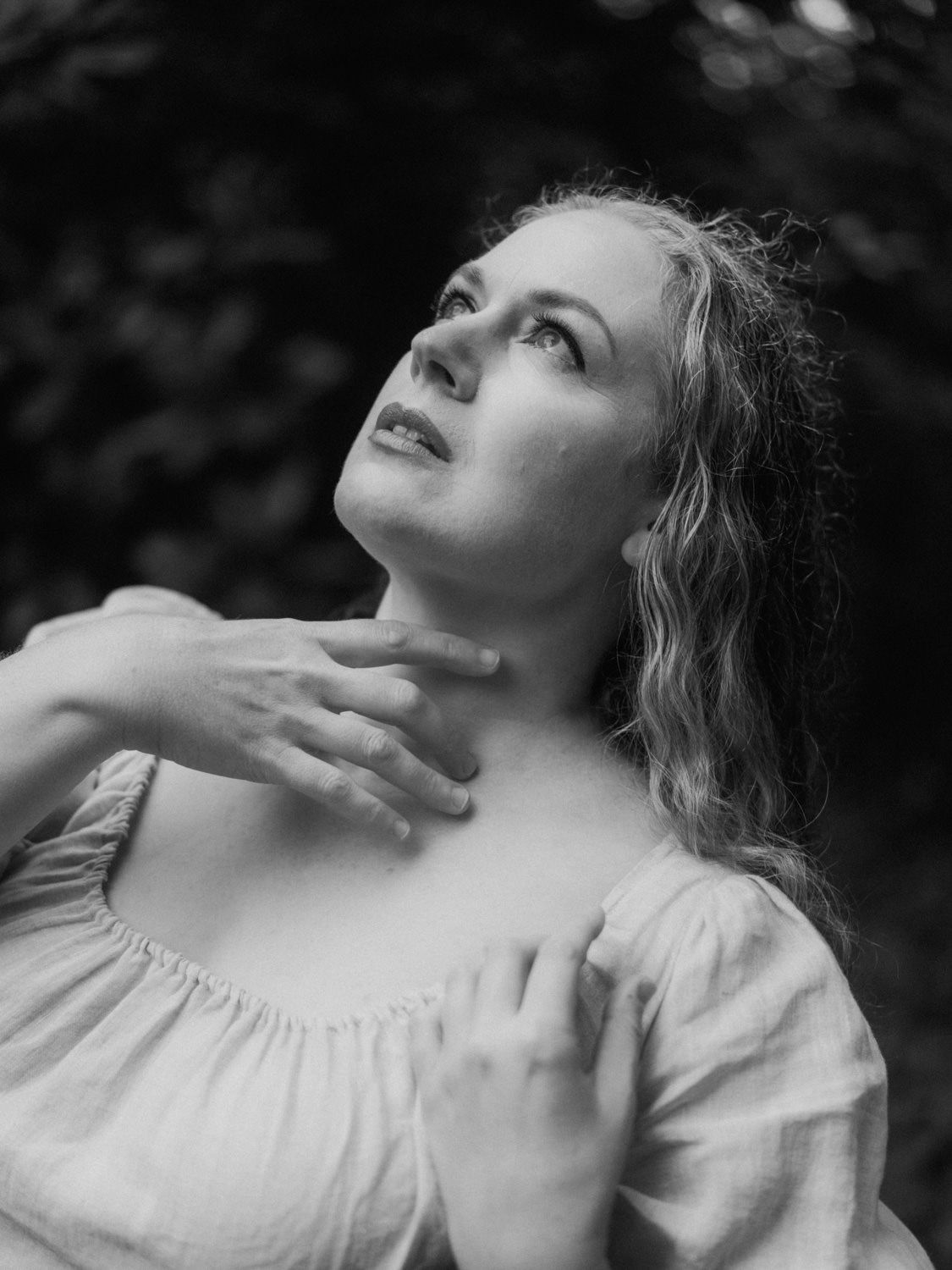 Black and white portrait of a young woman in a white dress looking upward, hand on chest, surrounded by nature.