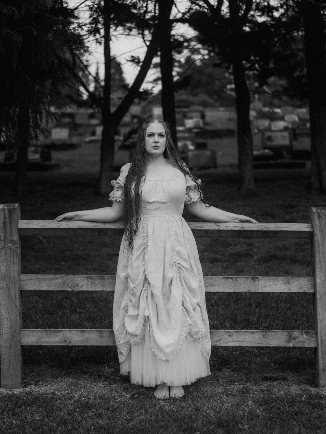 Black and white photo of a woman in a vintage white gown leaning on a wooden fence in a cemetery.