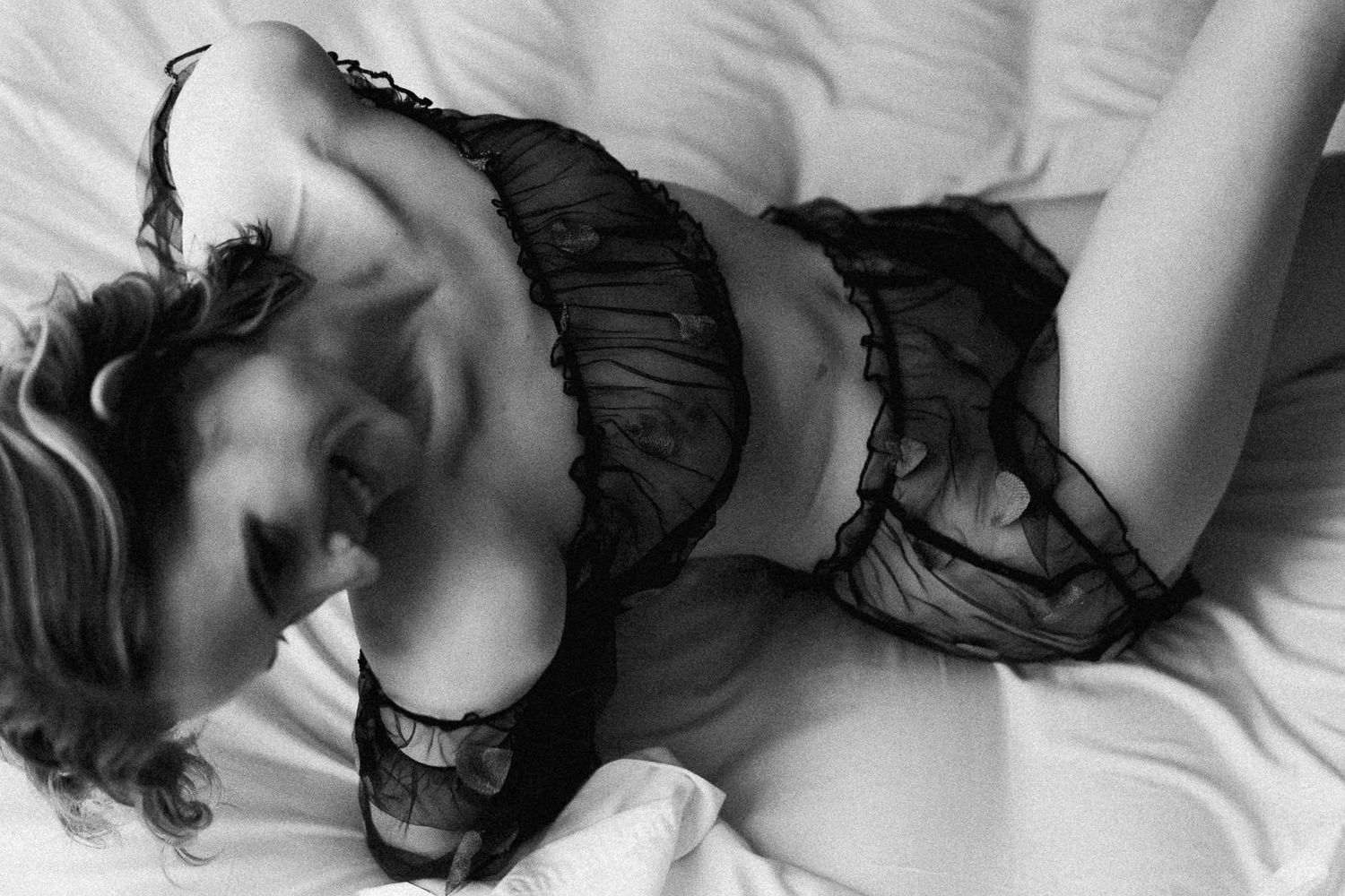 Black and white boudoir photo of a woman lying on white sheets wearing sheer lace lingerie.