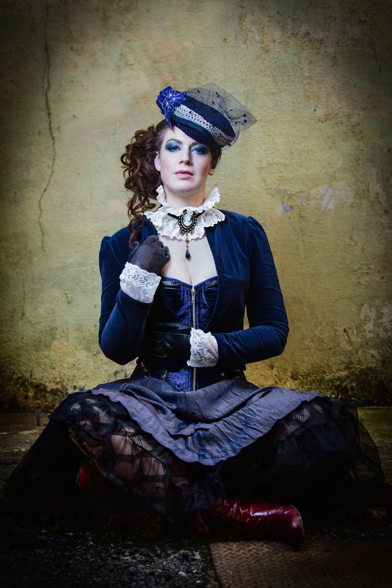 Woman in steampunk costume with blue velvet corset, lace details, mini hat, and red boots sits against aged wall.
