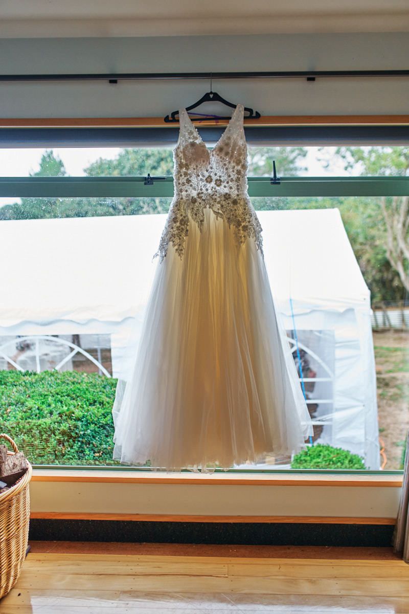 Elegant beaded wedding dress hanging on a hanger in front of a large window with garden views.