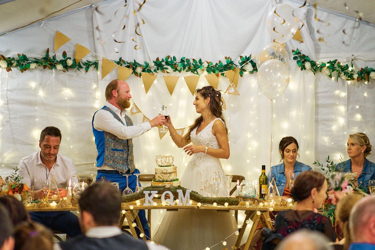 Bride and groom toast at wedding reception in decorated tent with fairy lights and bunting.