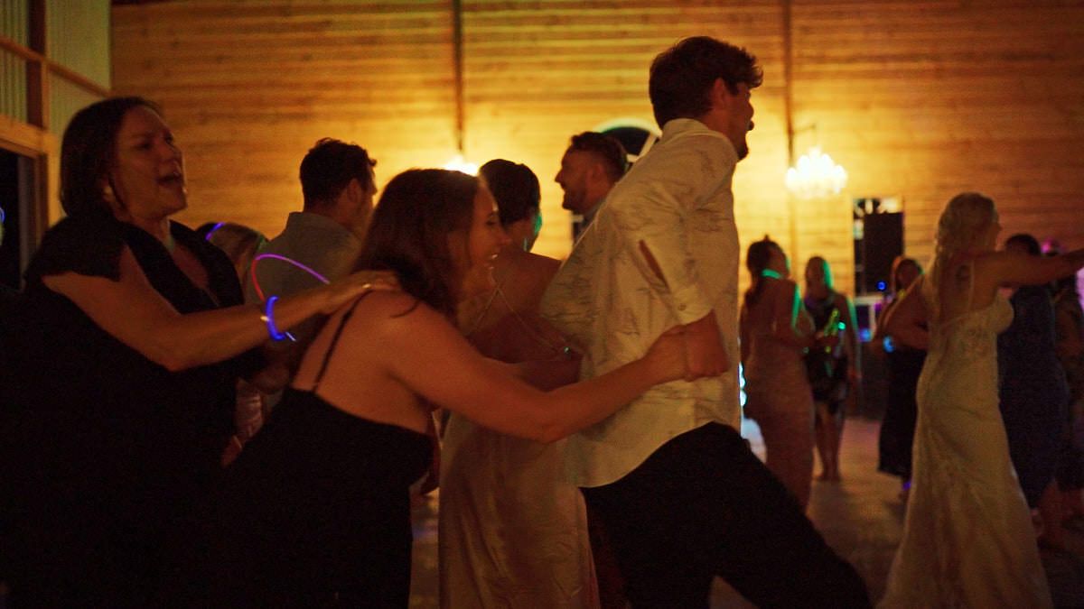 Guests dancing joyfully at a wedding reception in a warmly lit rustic venue with glowing dance floor lights.