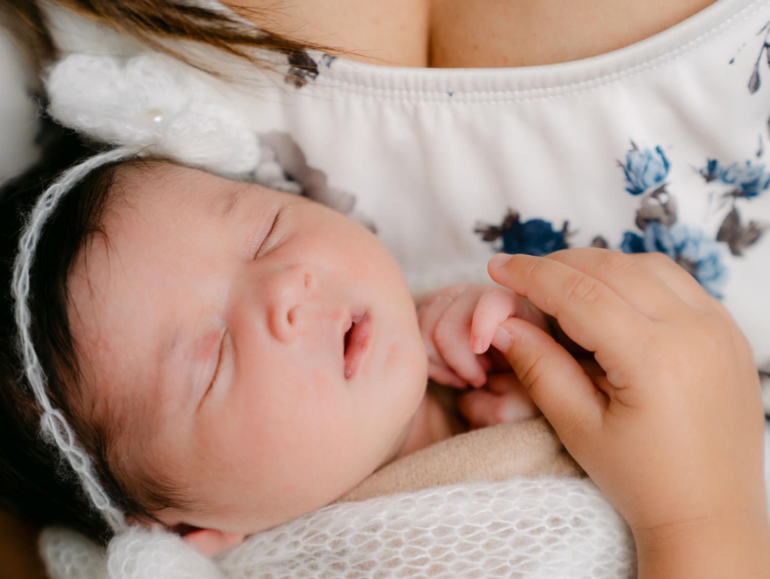 Newborn baby sleeping peacefully in mother's arms, wearing a white headband, wrapped in a soft white blanket.