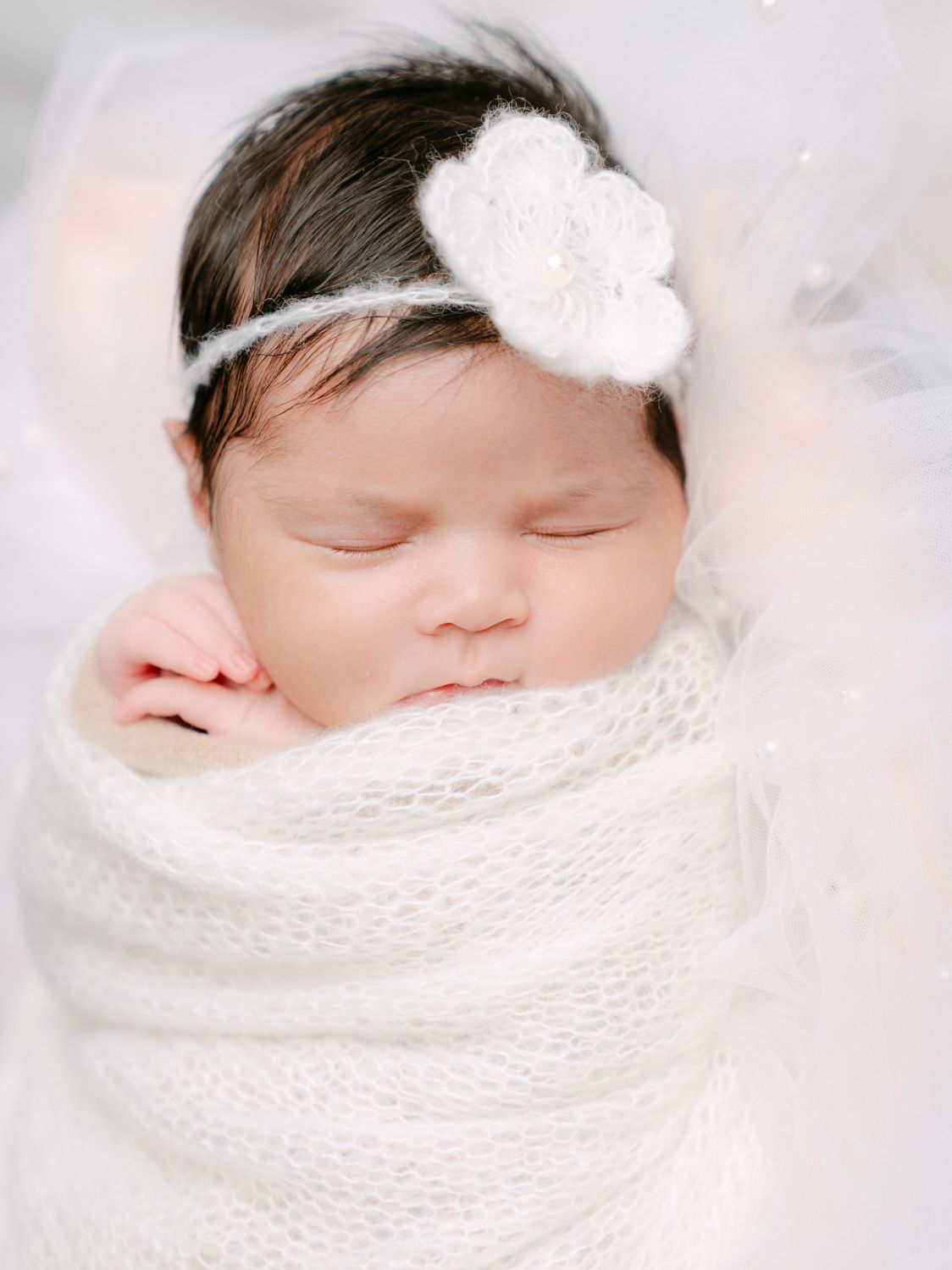Peaceful newborn baby girl swaddled in white knit wrap with crochet flower headband, sleeping serenely.