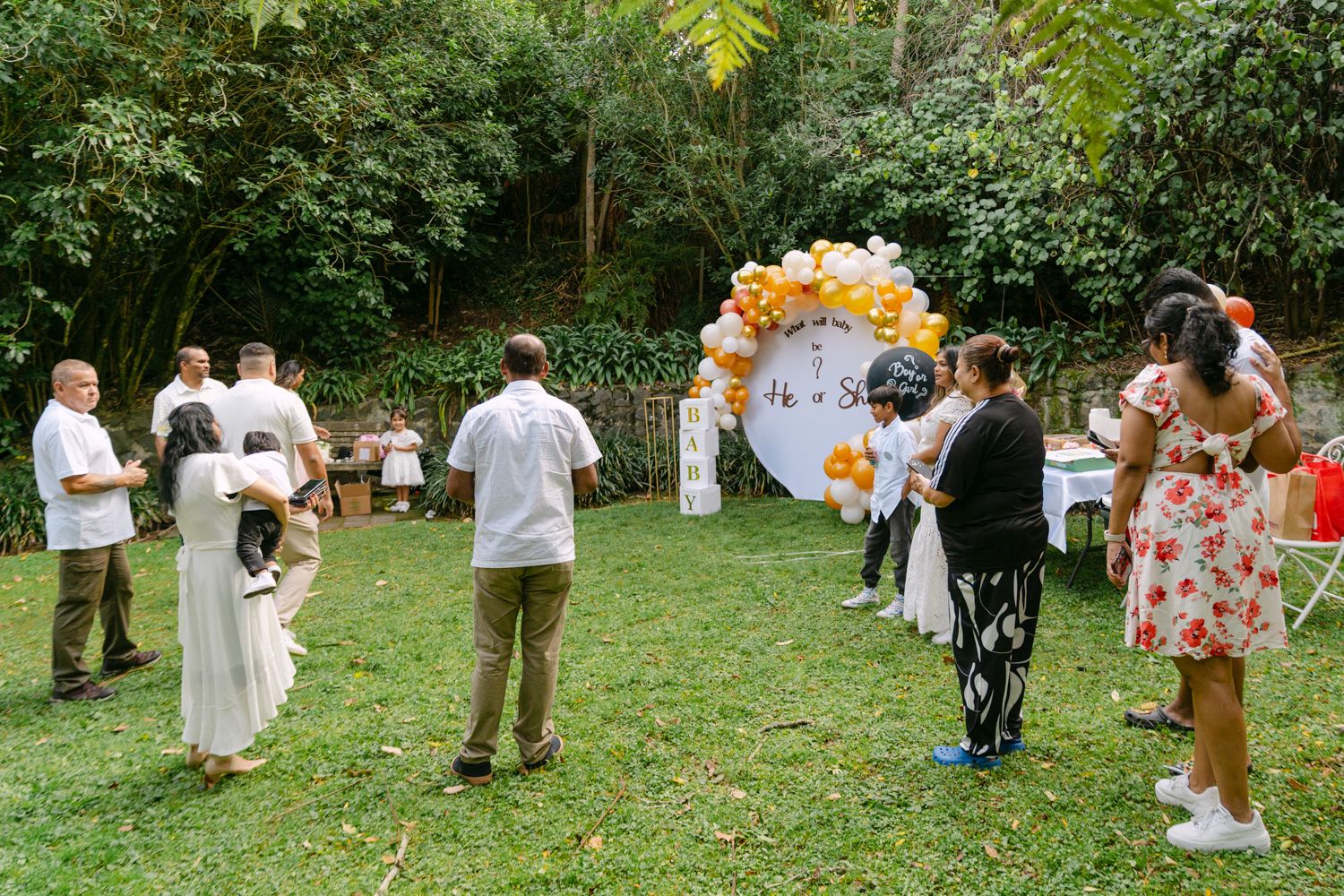 Outdoor garden party with guests gathered around a balloon arch backdrop on a lush green lawn.