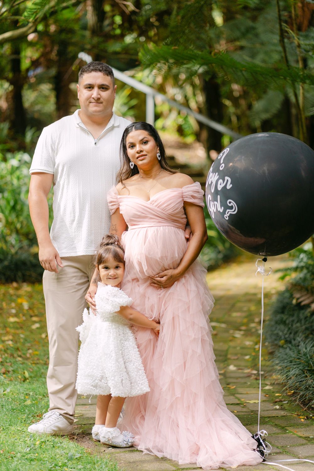 Family gender reveal party with pregnant woman in pink gown, man in white shirt, and young girl holding black balloon outdoors.