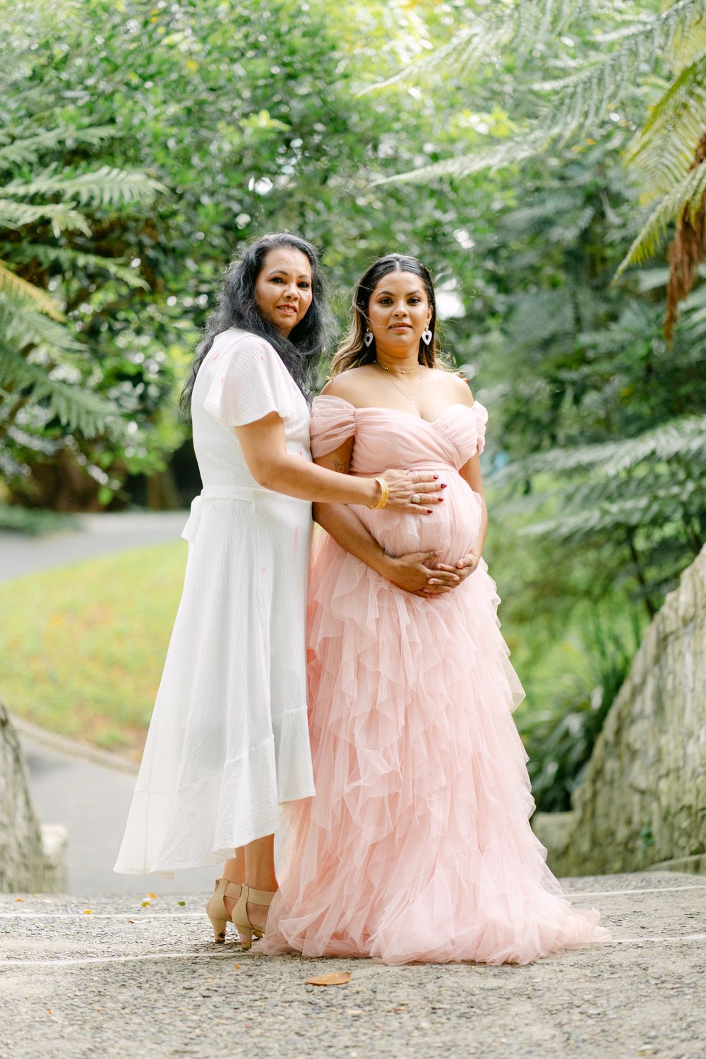 Two women pose outdoors, one in white dress, other in pink ruffled maternity gown, surrounded by lush greenery.