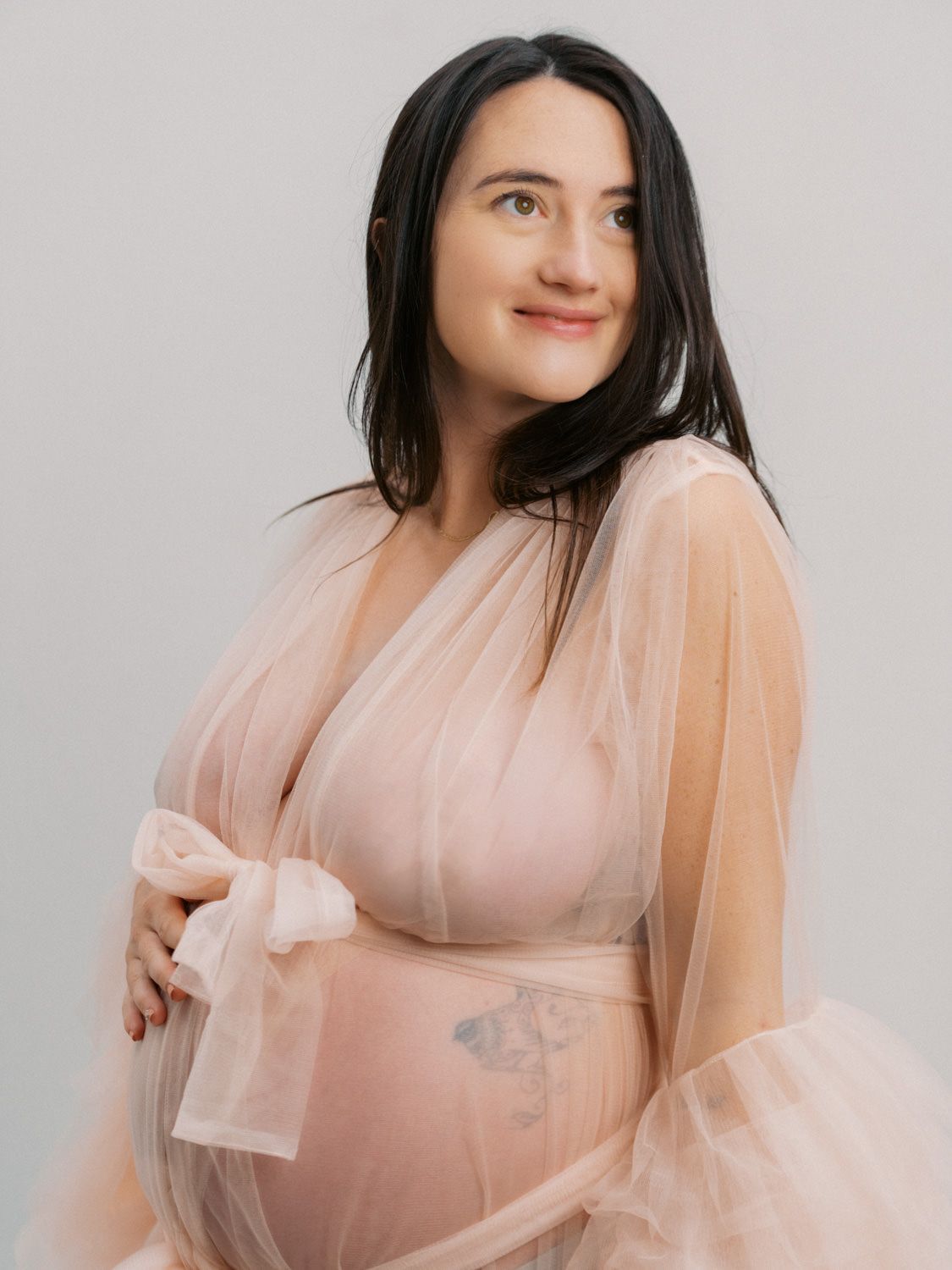 Pregnant woman in sheer blush pink tulle maternity gown cradling baby bump in studio portrait.