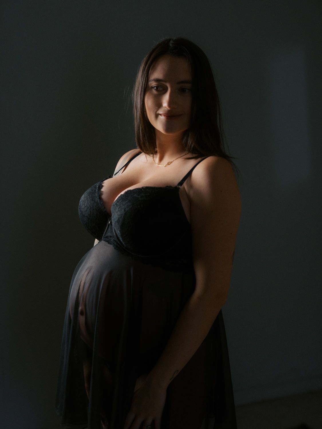 Pregnant woman in black lingerie and sheer skirt posing in dramatic studio lighting for maternity portrait.