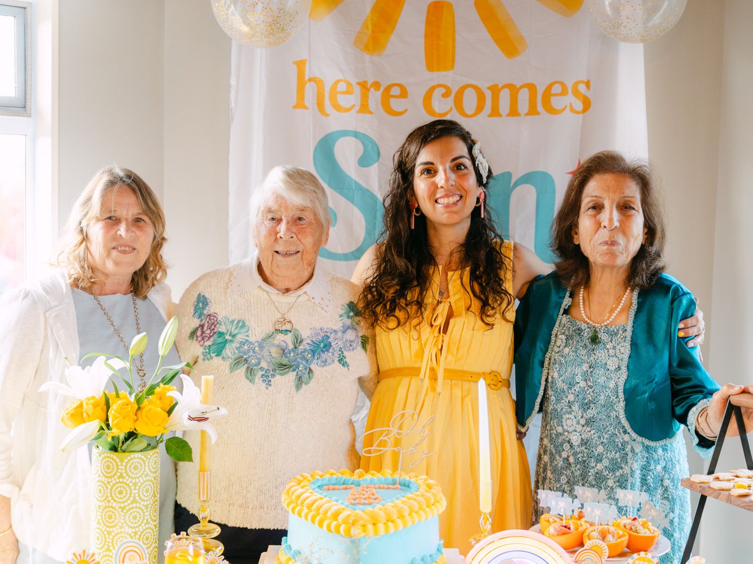 Four women pose together at a sunshine-themed baby shower with a cake and 'Here Comes Sun' banner.