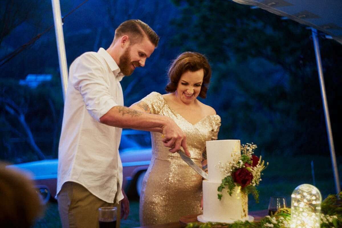 Couple cutting white wedding cake at outdoor evening reception, bride in gold dress, groom in white shirt.