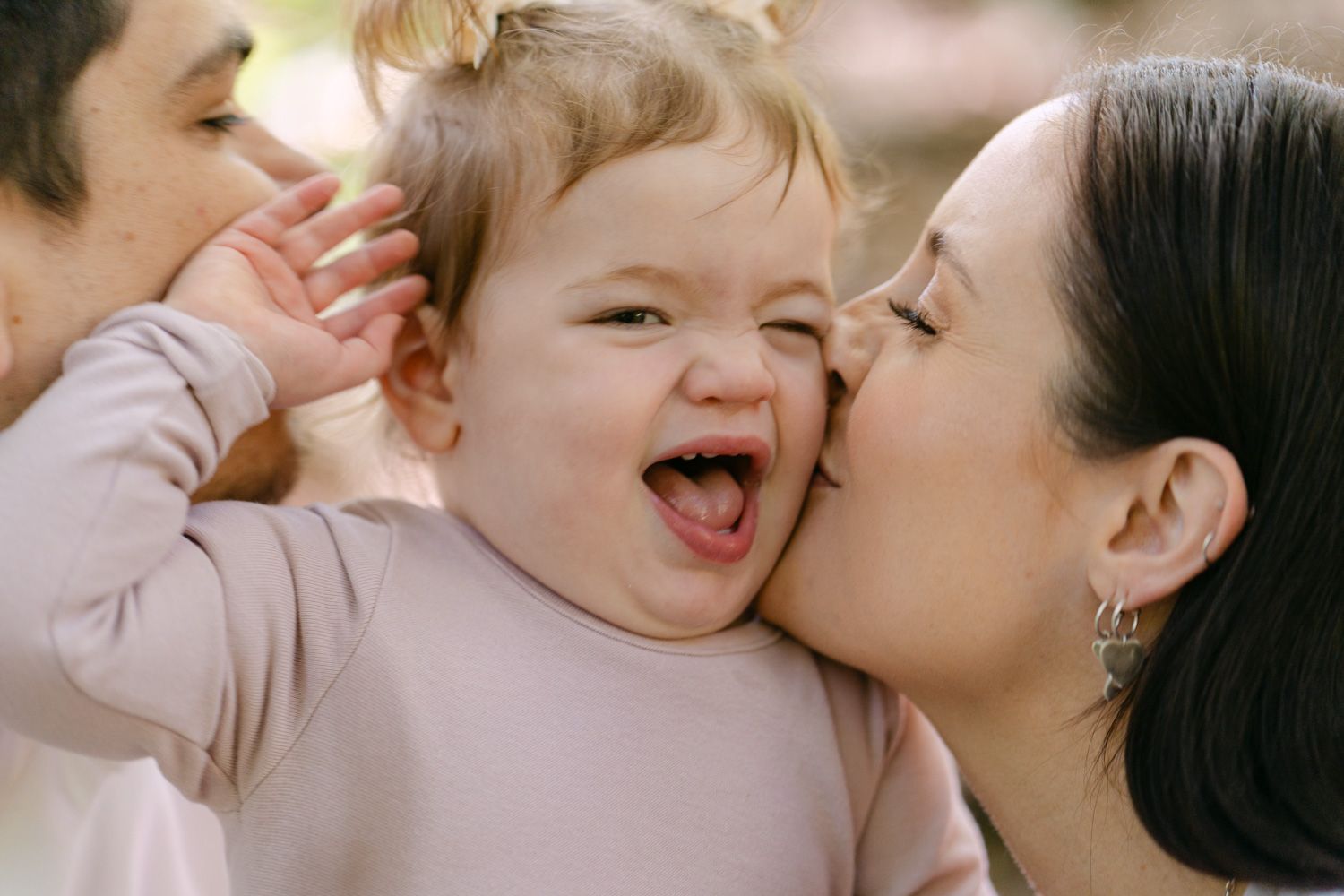 Happy baby laughing while parents kiss cheeks in a sweet outdoor family portrait moment.