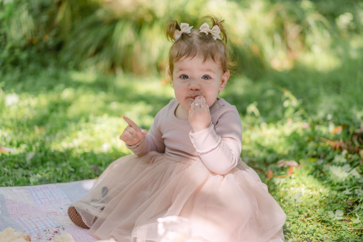 A baby girl in a pink tutu dress and floral headband sitting on grass at an outdoor birthday party.
