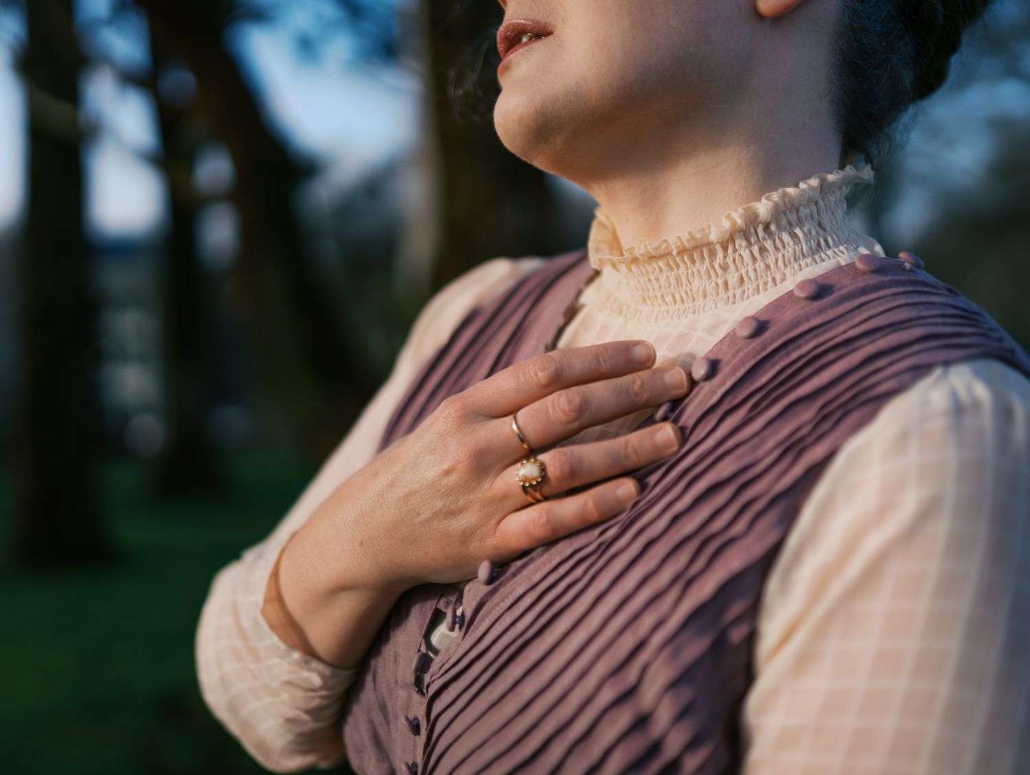 Woman in vintage dress with hand on chest wearing gold ring, standing outdoors near trees in soft light.