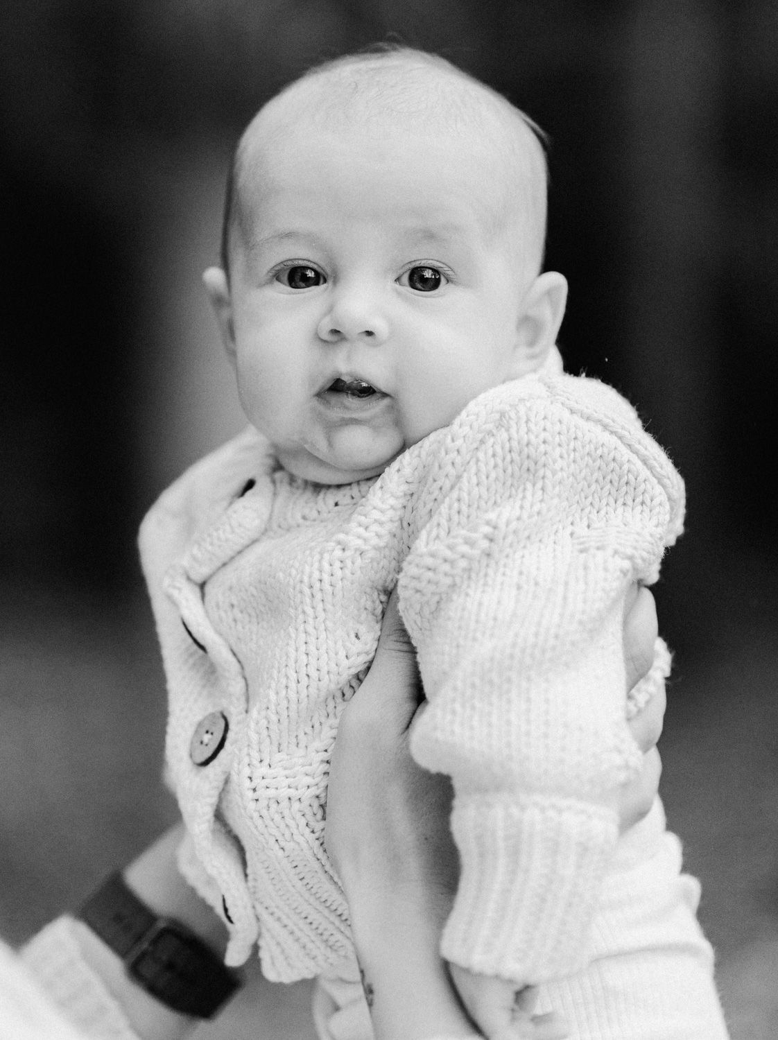 Black and white photo of an adorable baby in a knit sweater being held up by an adult's hands.