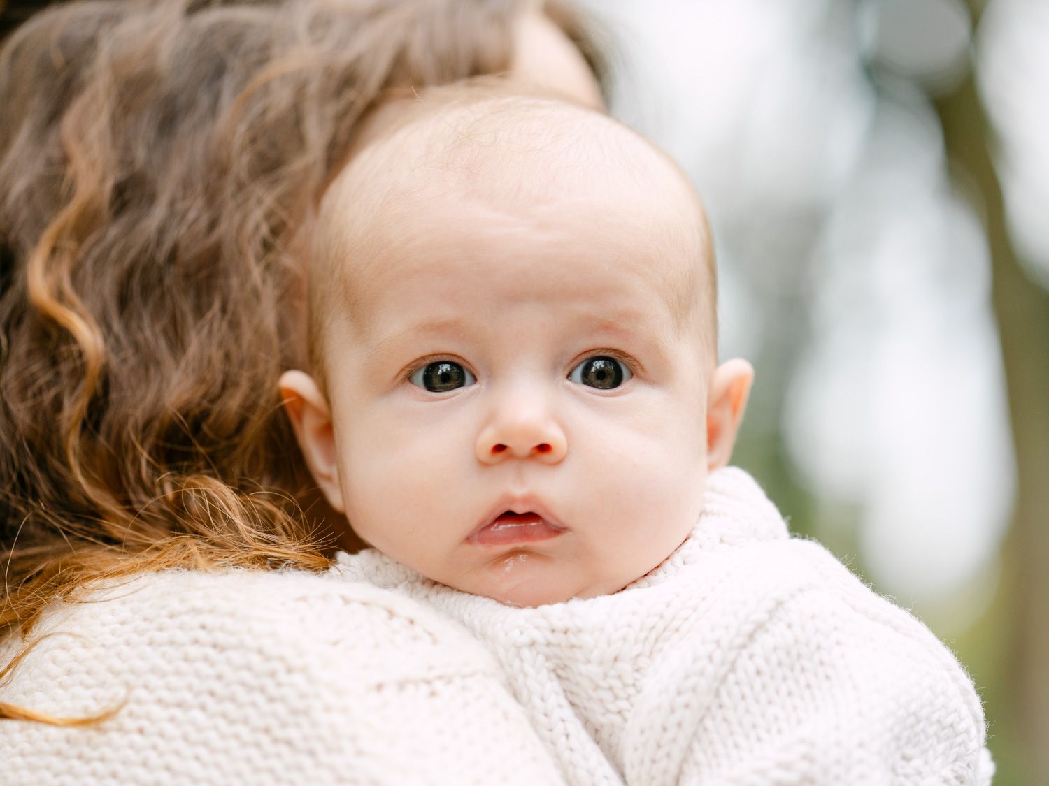 A curious baby with wide eyes rests on a mother's shoulder outdoors, wearing a white knit sweater.