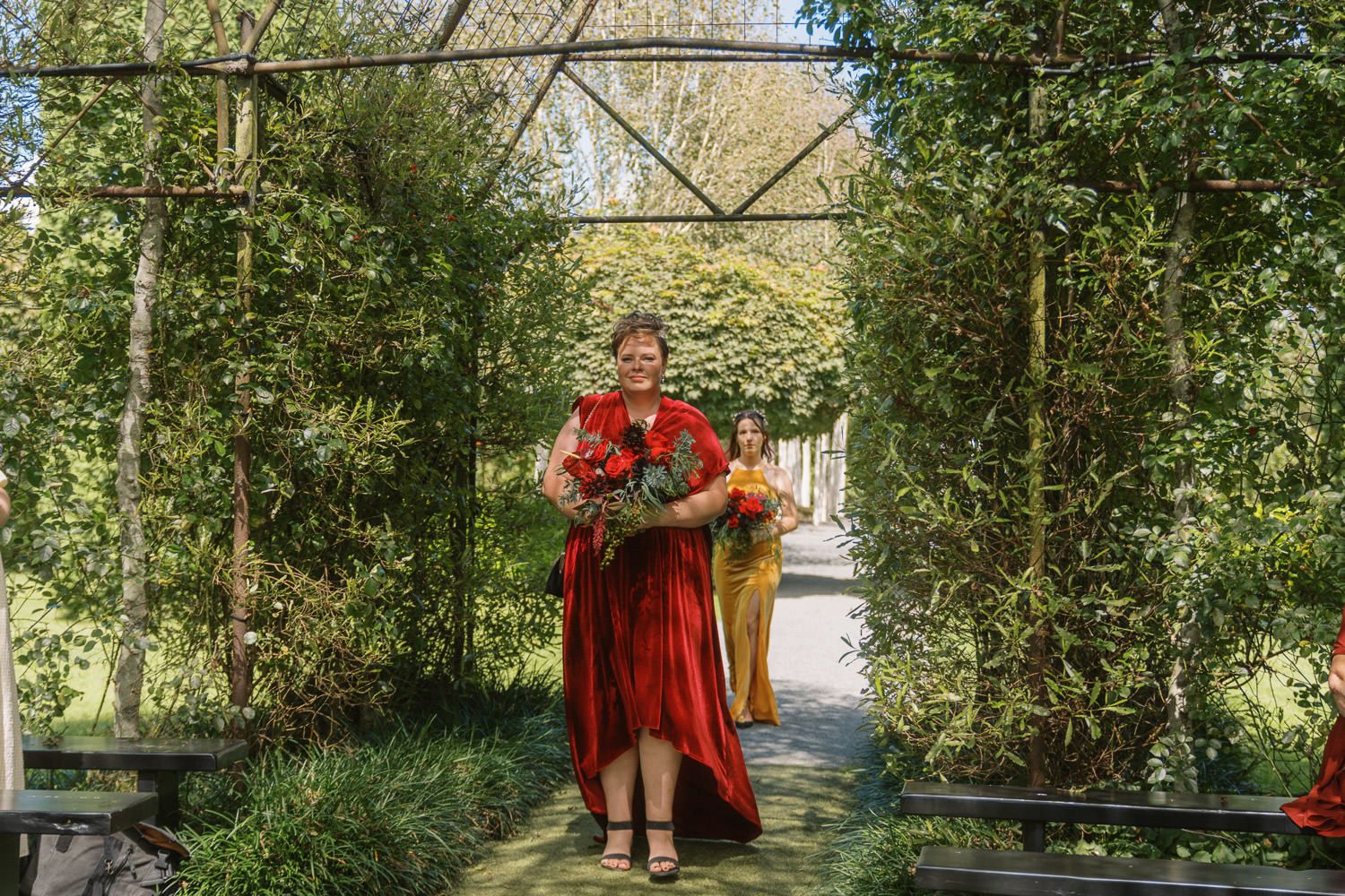 Woman in red dress walks through green garden archway holding colorful floral bouquet on sunny day.