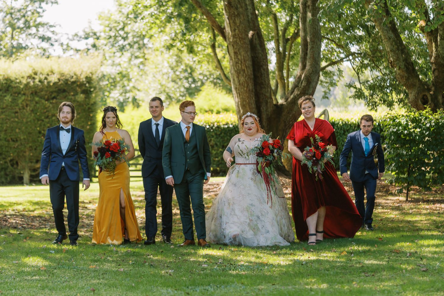 Wedding party posing outdoors in colorful formal attire including gold, burgundy, and teal with red bouquets.