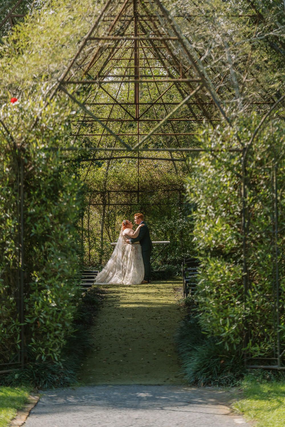 Wedding couple shares romantic moment under elegant metal pergola arch framed by vibrant green garden foliage.