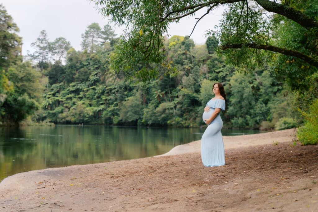 Pregnant woman in light blue dress stands peacefully by a serene river surrounded by lush green trees.