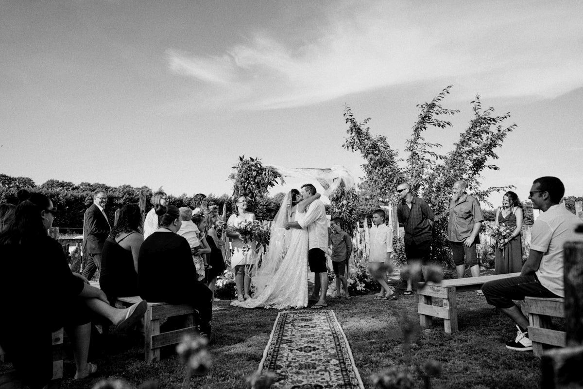 Black and white outdoor wedding ceremony with couple at altar surrounded by guests seated on benches in a garden setting.